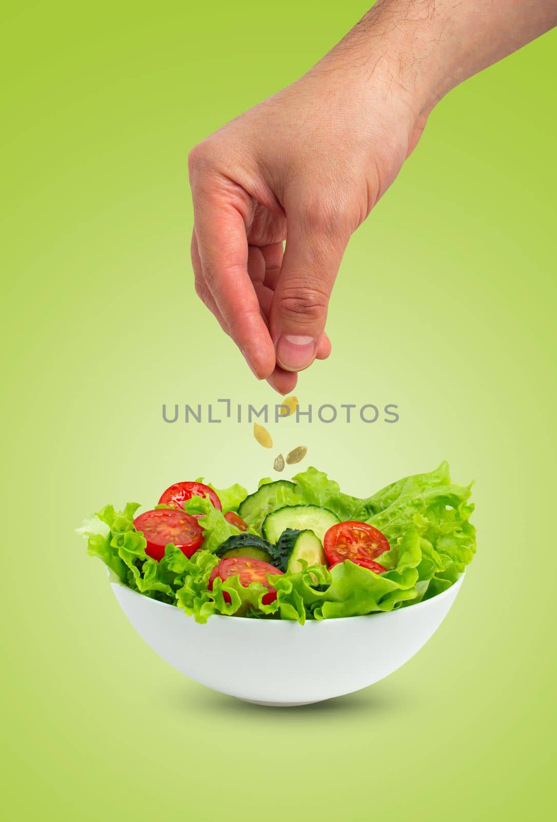 White bowl of salad with fresh cucumbers, tomatoes, iceberg salad leaves and male hand pouring oil with spoon over salad isolated on green background