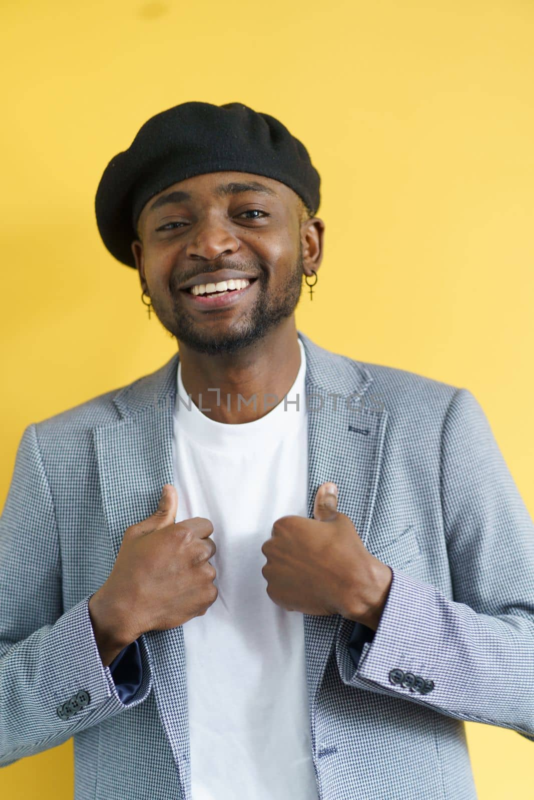 Happy stylish african american man smiling on yellow background. Young student with earrings in suit and beret posing for camera. by LipikStockMedia