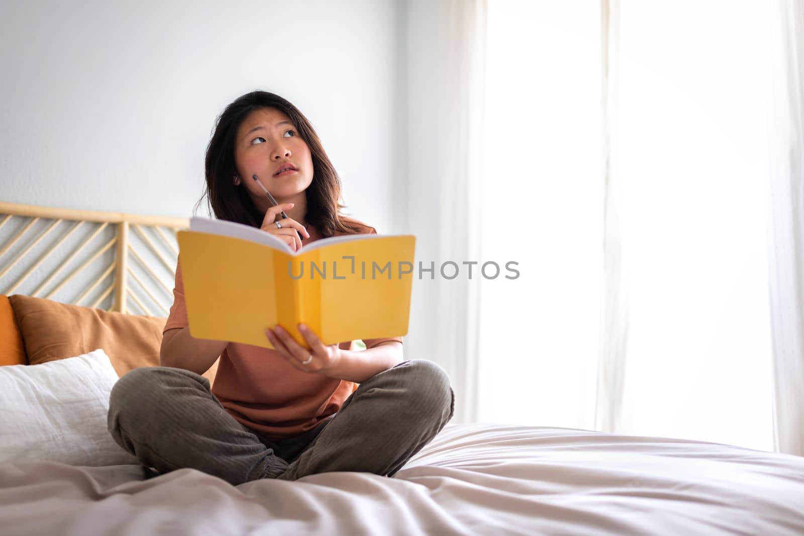 Teen asian girl sitting on bed thinking, using diary and pen to write down thoughts and ideas. Copy space. Lifestyle concept.