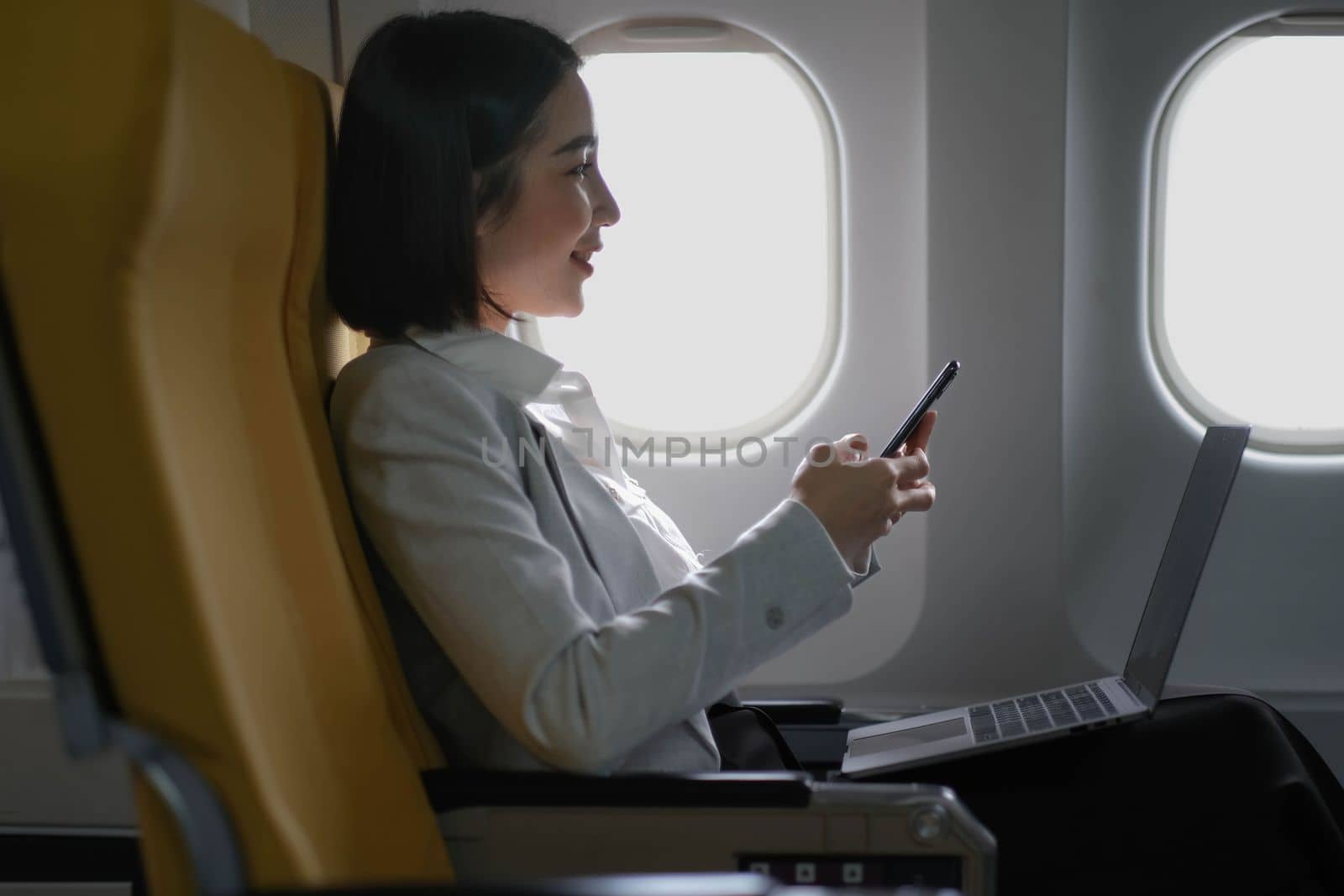 Asian woman passenger sitting in airplane near window and reading news from social networks or using travel applications in smartphone.