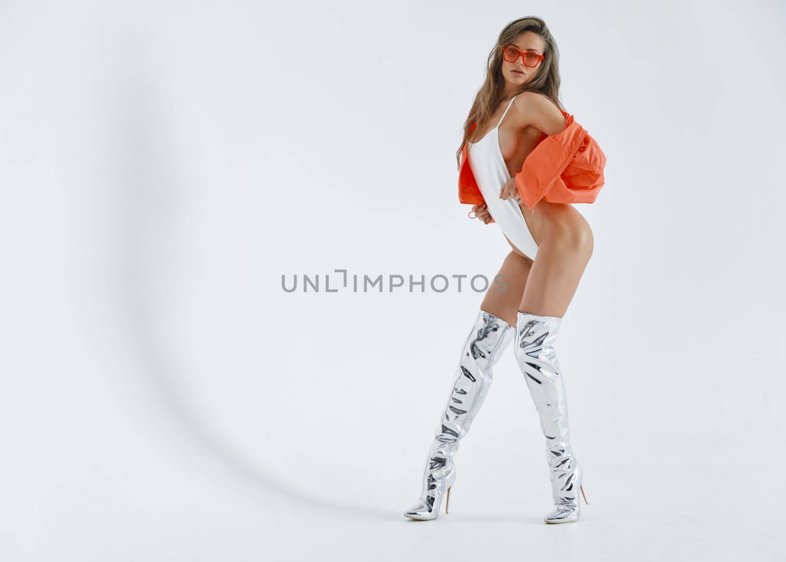 A beautiful girl of an athletic physique erotic moves in front of the camera, she is dressed in high boots of silver color, white bodysuit, pink jacket and glasses, long brown hair, white background. High quality photo