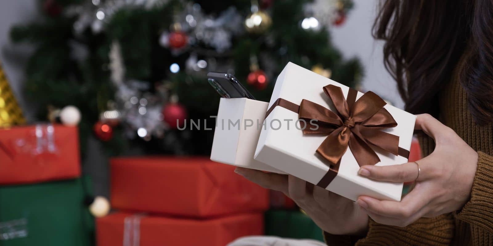 Smiling asian woman in festive mood holding wrapped gift box with brown ribbon in hands, stretching out present to camera, celebrating Merry Christmas