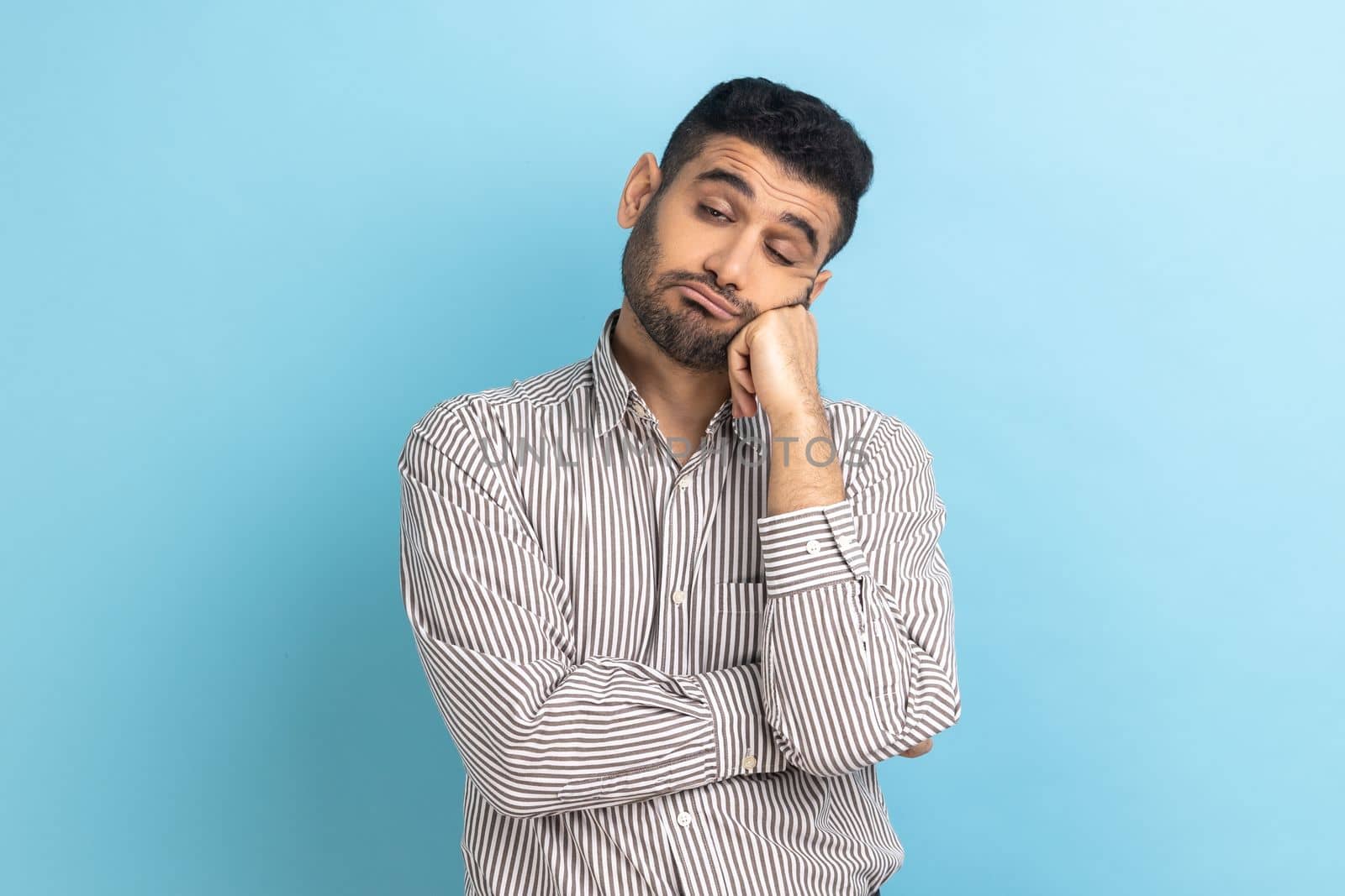 Portrait of sad upset or bored bearded businessman standing and looking at camera with dissatisfied sadness face, wearing striped shirt. Indoor studio shot isolated on blue background.