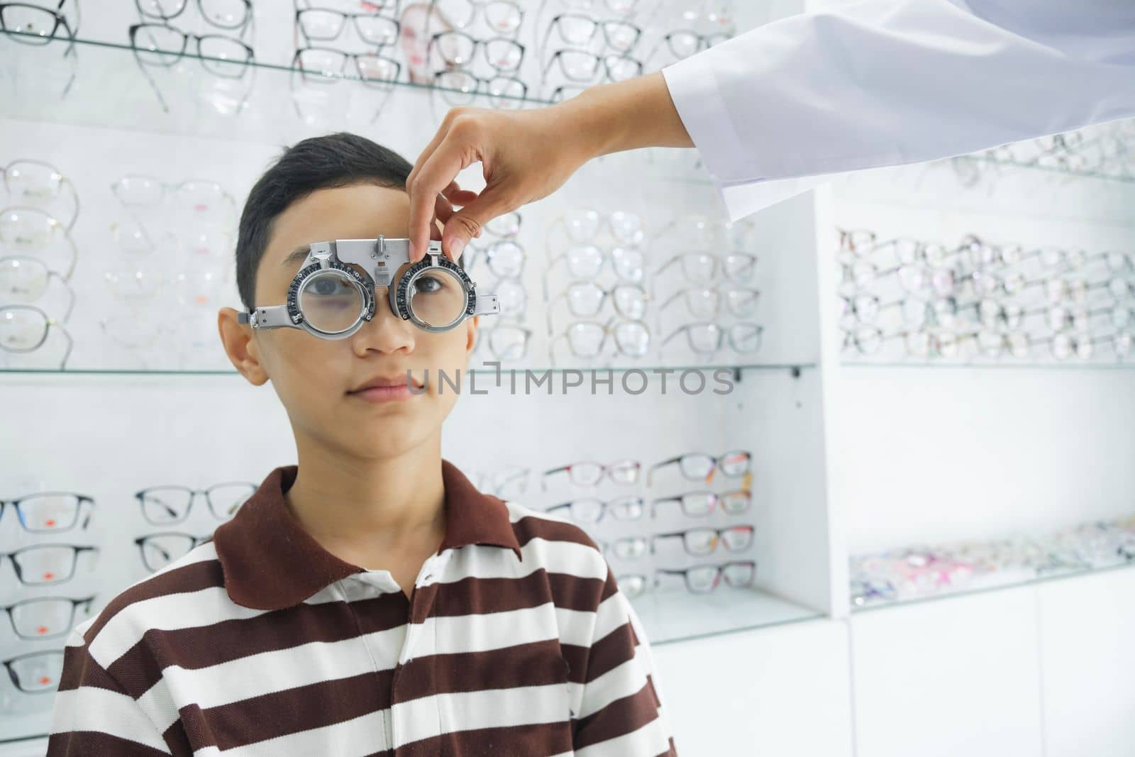 Smart young boy sitting in optometrist cabinet having his eyesight checking, examining, testing with trial frame glasses by professional optician for new pairs of eyeglasses.