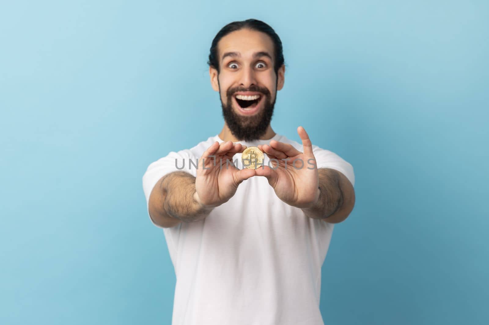 Portrait of surprised man with beard wearing white T-shirt standing holding out bitcoin, paying attention at new digital cryptocurrency. Indoor studio shot isolated on blue background.