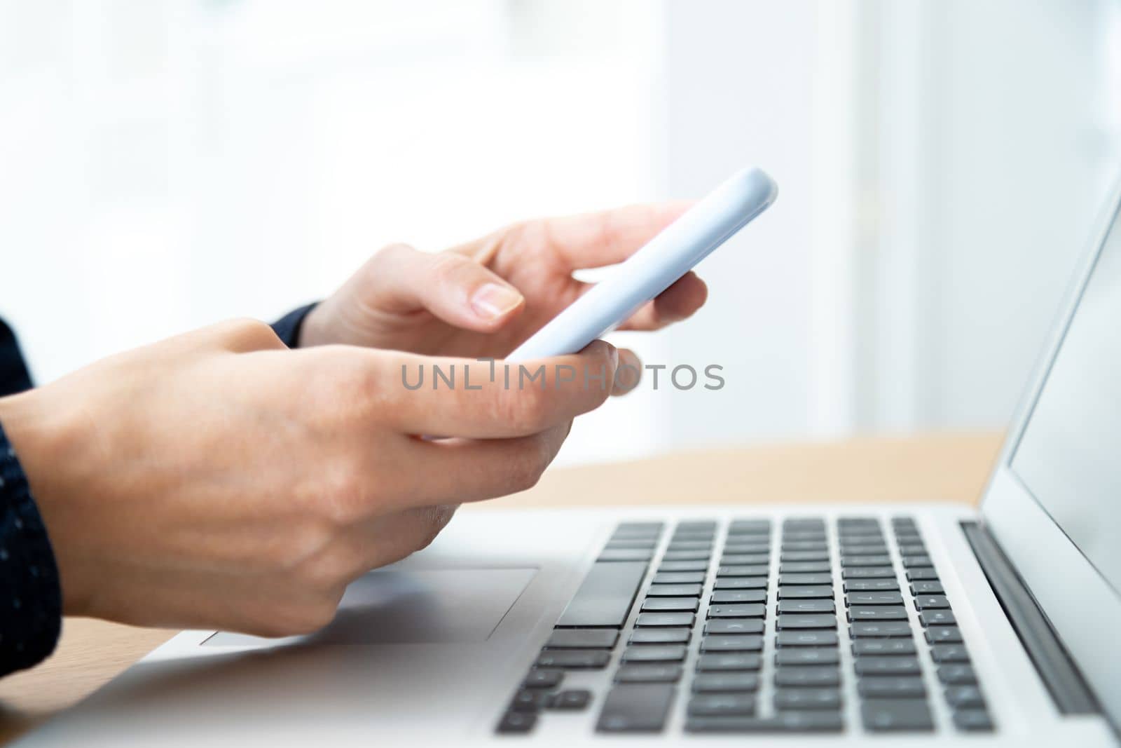Close-up image of woman hands using mobile phone while working in the office with laptop. Selective focus on hands. Blurred background. Technology concept.