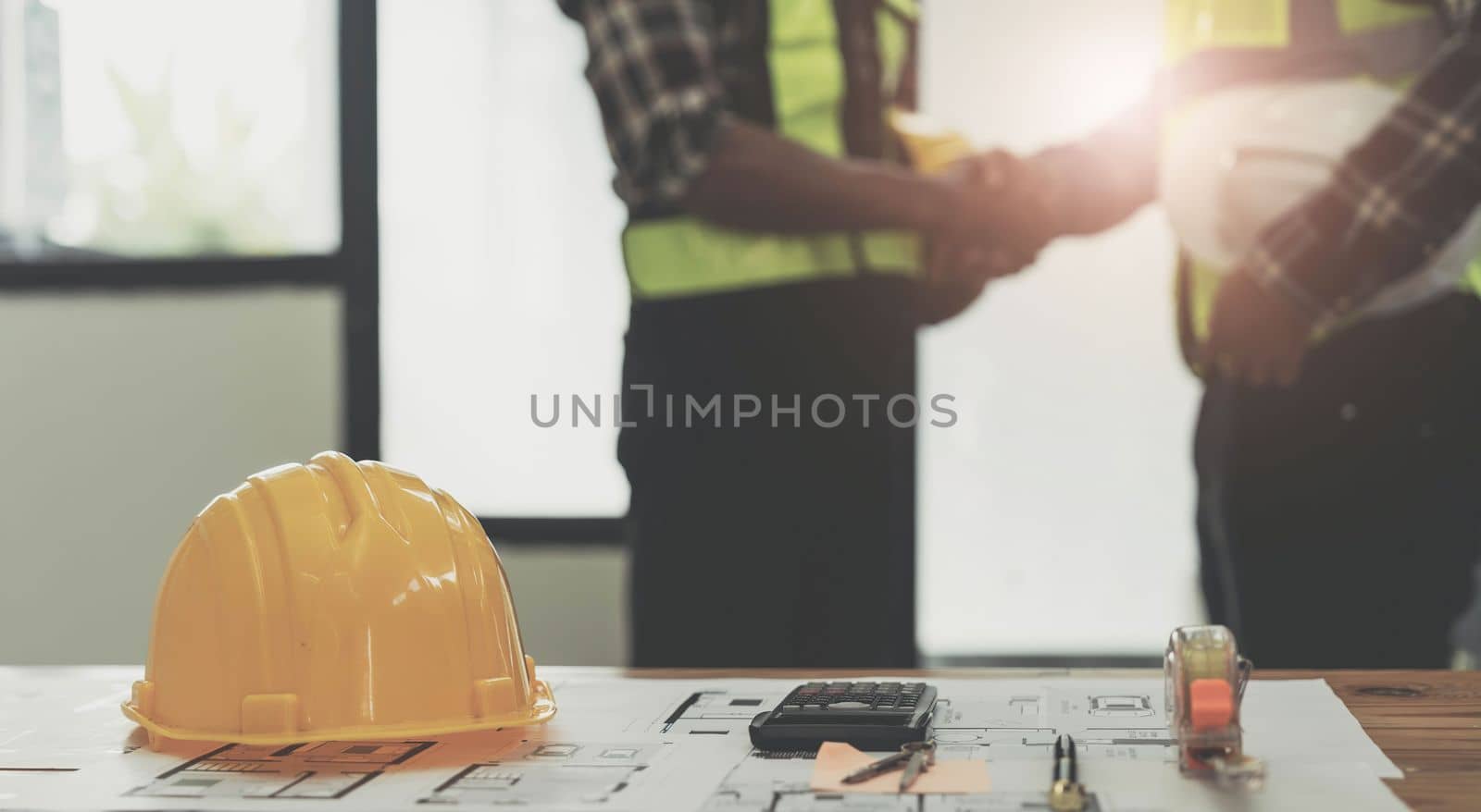yellow safety helmet on workplace desk with construction worker team hands shaking greeting start up plan new project contract in office center at construction site, partnership and contractor concept..