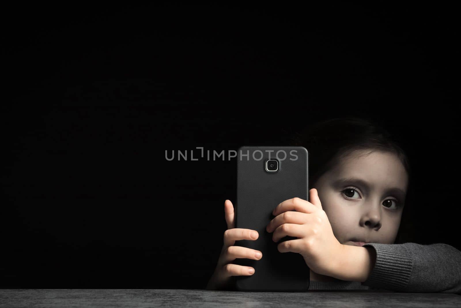 Little child with a gray face hides behind a large smartphone in his hands, on a black background. Kid gadget addiction and insomnia, psychological problems, copy space