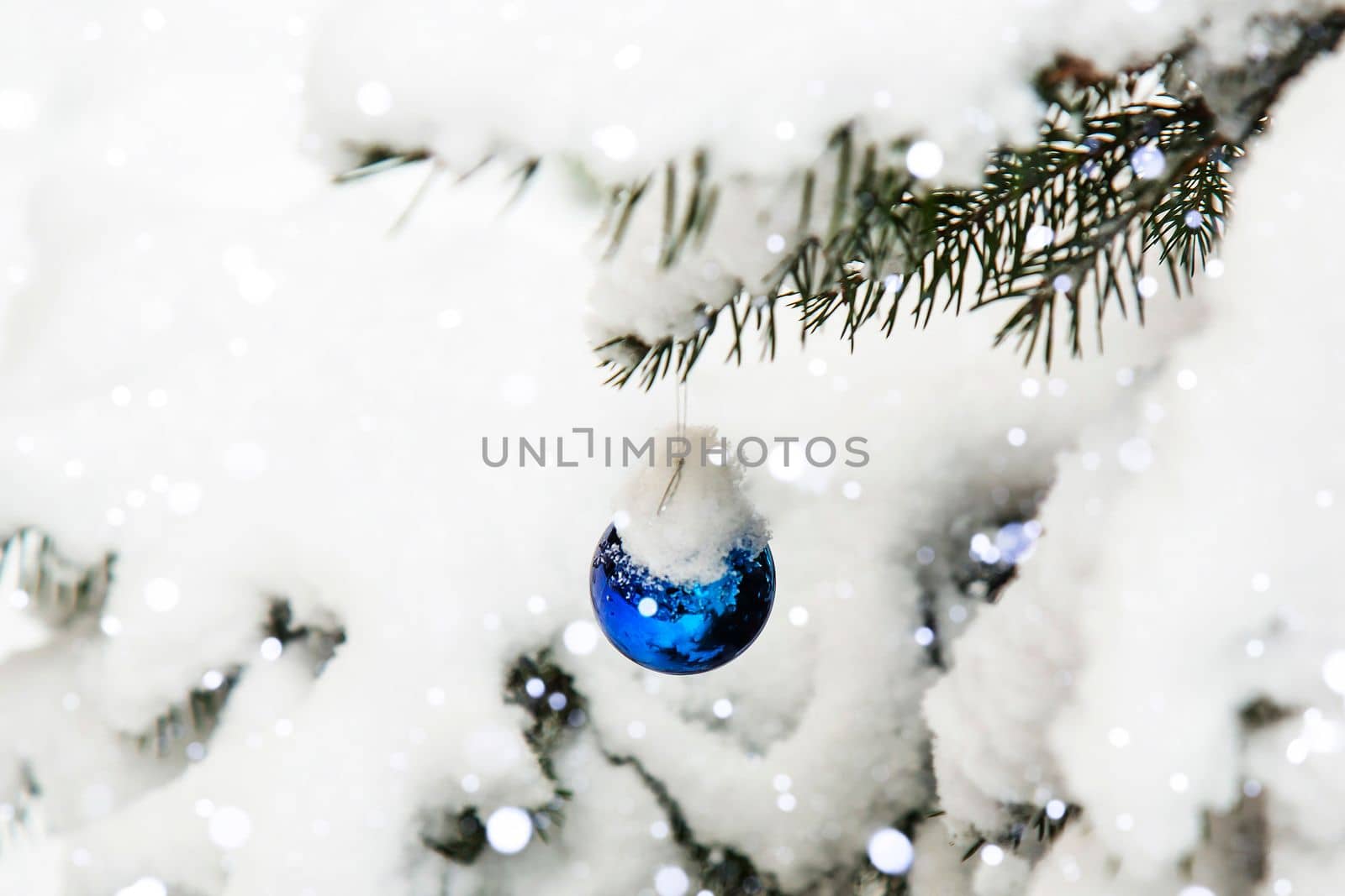 A branch of a Christmas tree in the snow with a Christmas decoration. A blue Christmas toy is hanging on a tree outside. Snowfall. Lots of snow. Winter, festive background.