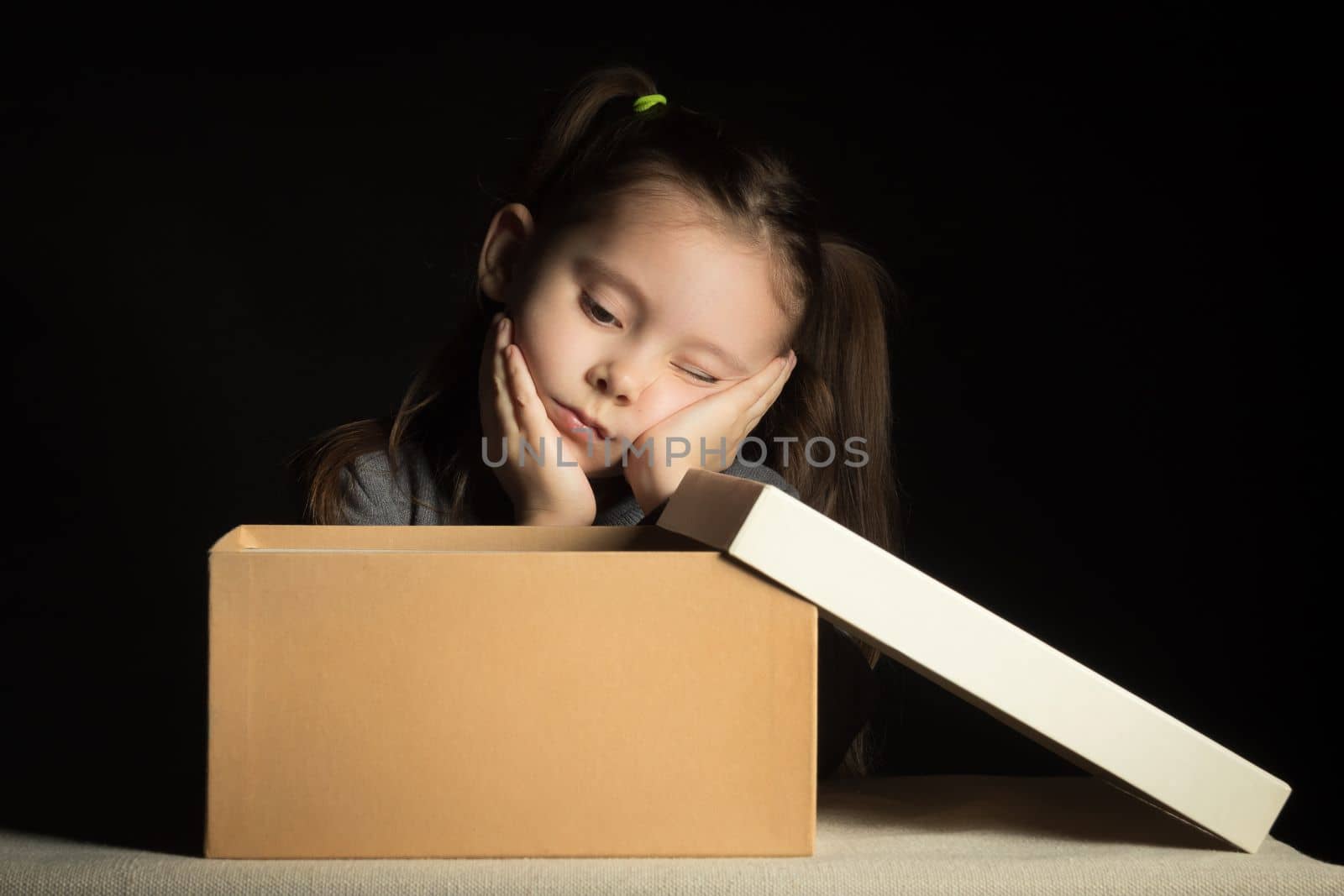 Upset and disappointed little girl opens a long-awaited parcel with a gift, copy space on cardboard box.