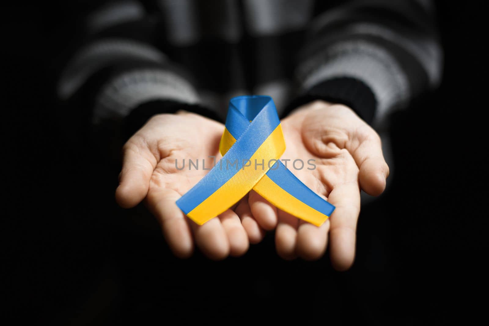 childs hand holds blue and yellow ukraine ribbon on dark background. concept needs help and support, truth will win