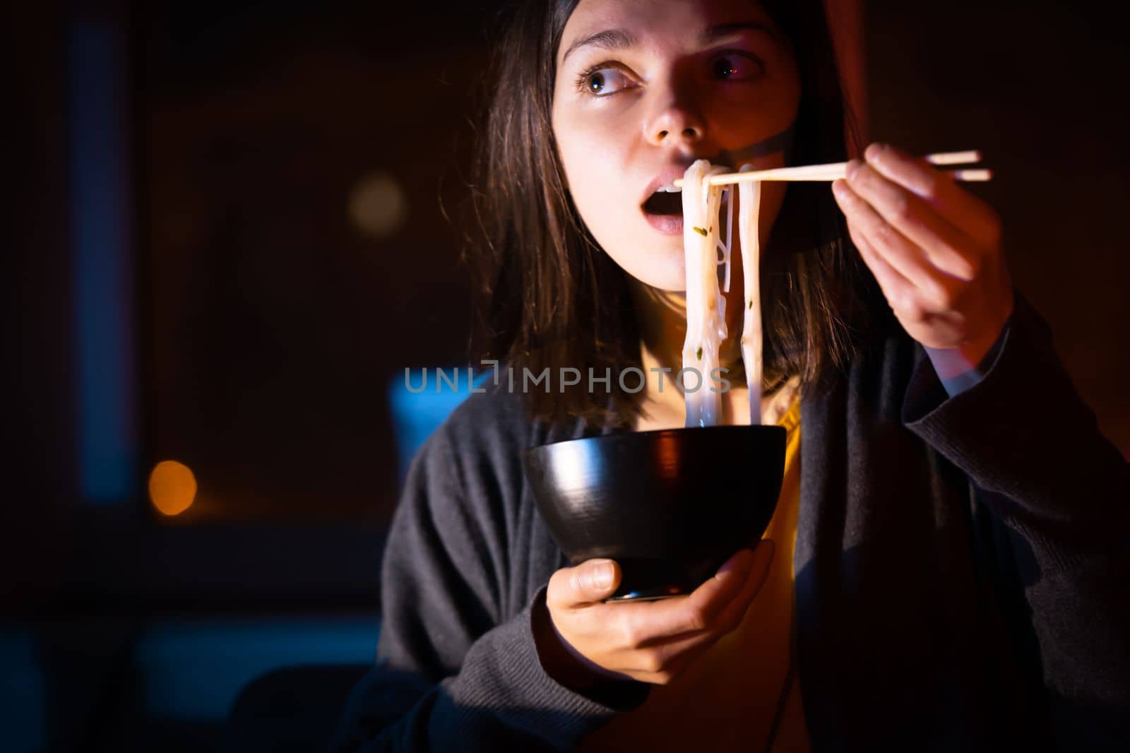 Young girl works at night at home with laptop and eats noodles, asian food with chopsticks. A woman watches a show, a movie and has dinner with ordered food from a cafe.