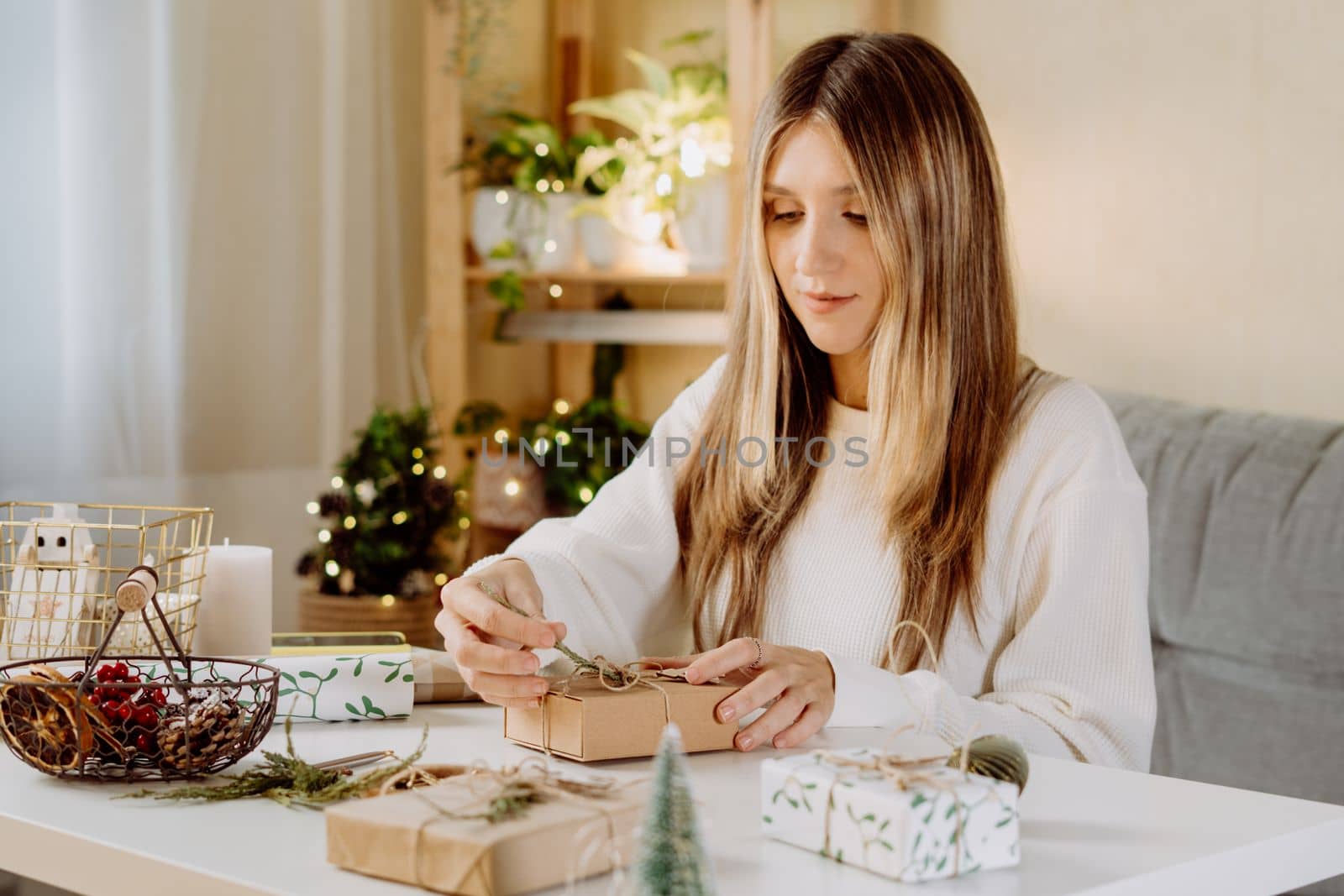 Woman wrapping Christmas eco natural gift boxes at home. Preparing presents on white table with decor elements, lights and items Christmas or New year DIY packing Concept.