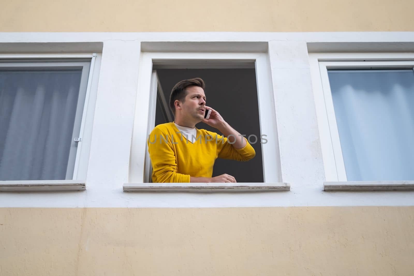 Young man standing near a window, looking outside and talking on his cellphone. Reporting a crime.