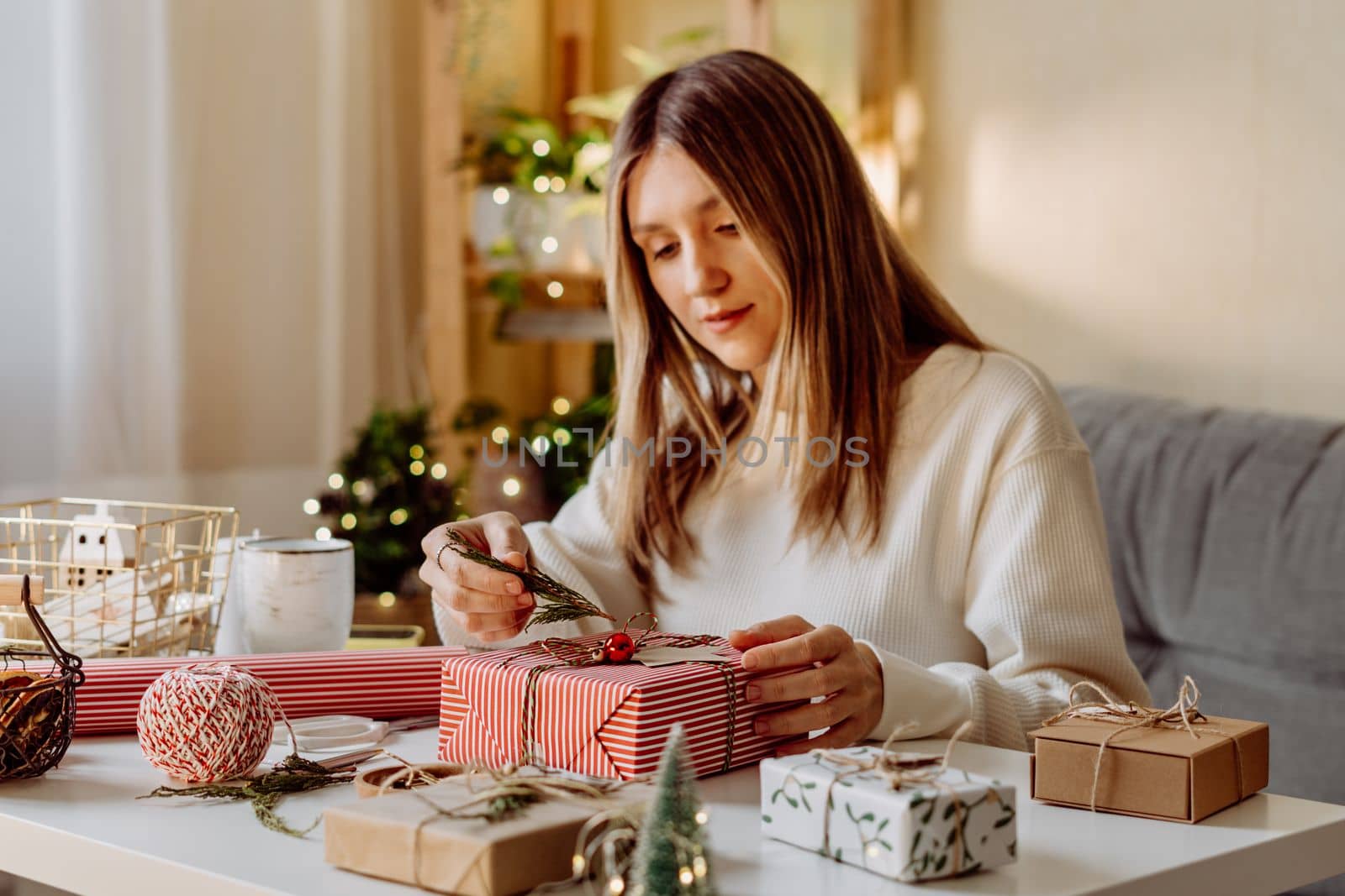 Woman wrapping Christmas eco natural gift boxes at home. Preparing presents on white table with decor elements, lights and items Christmas or New year DIY packing Concept.