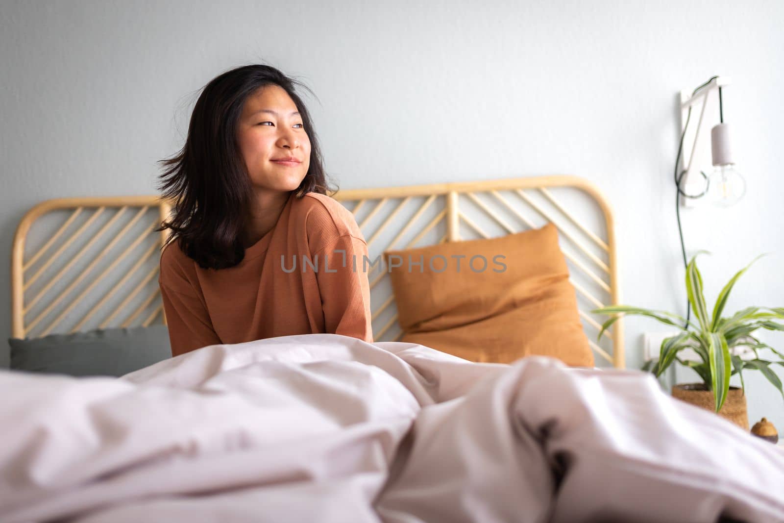 Happy young asian woman sitting on bed after waking up in the morning in beautiful, cozy bedroom at home. Lifestyle concept.