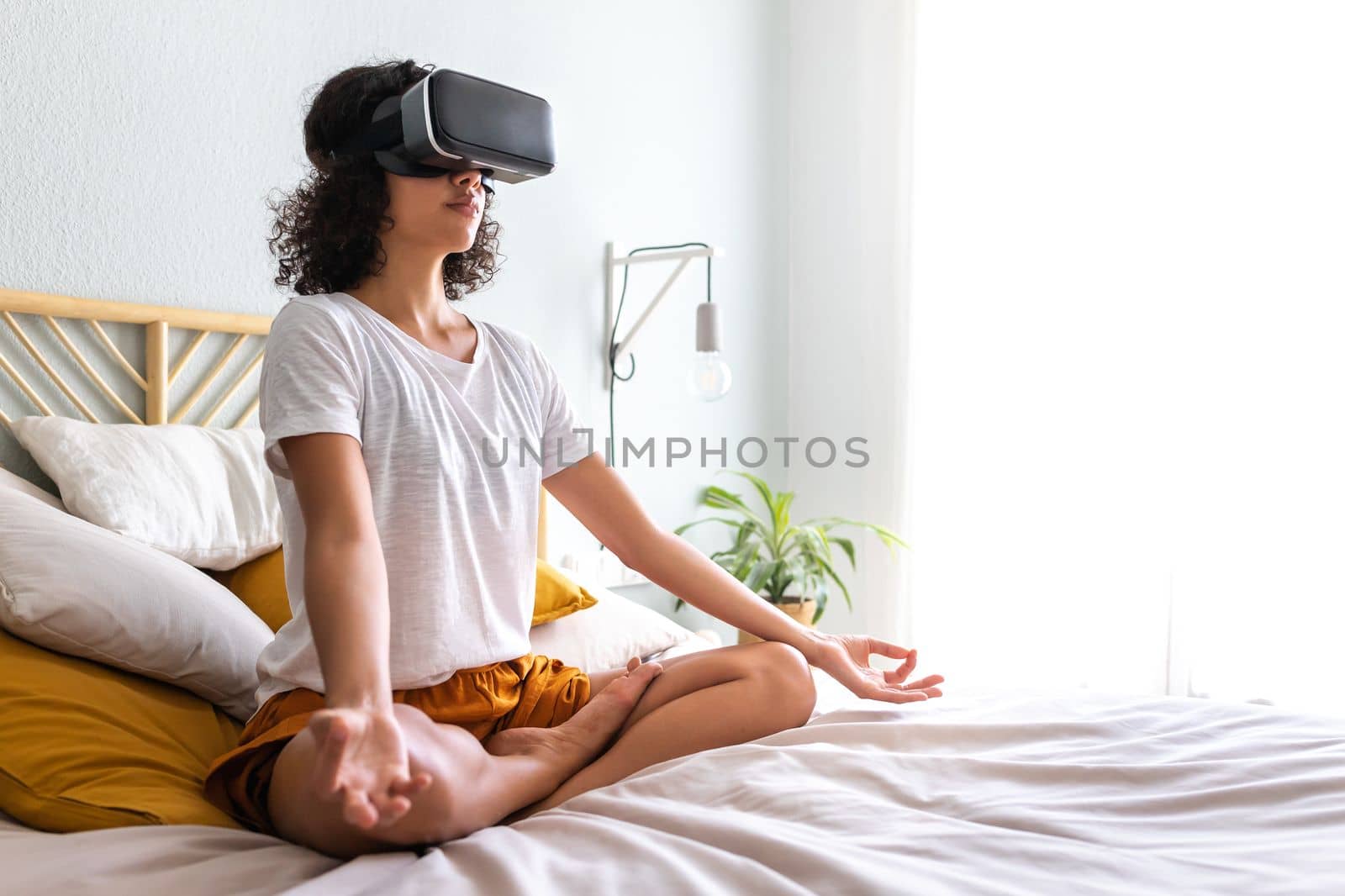 Young multiracial latina woman meditating with help of VR experience app sitting on bed. Hispanic girl doing meditation using virtual reality goggles at home. Copy space. Technology and lifestyle concepts.