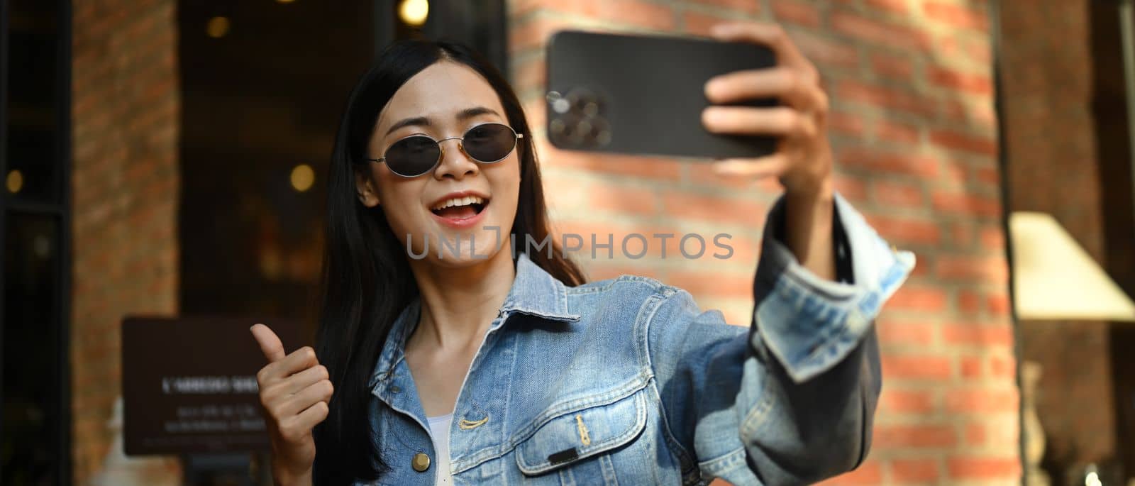 Cheerful stylish woman taking selfie or making video call on mobile phone while standing in city street.