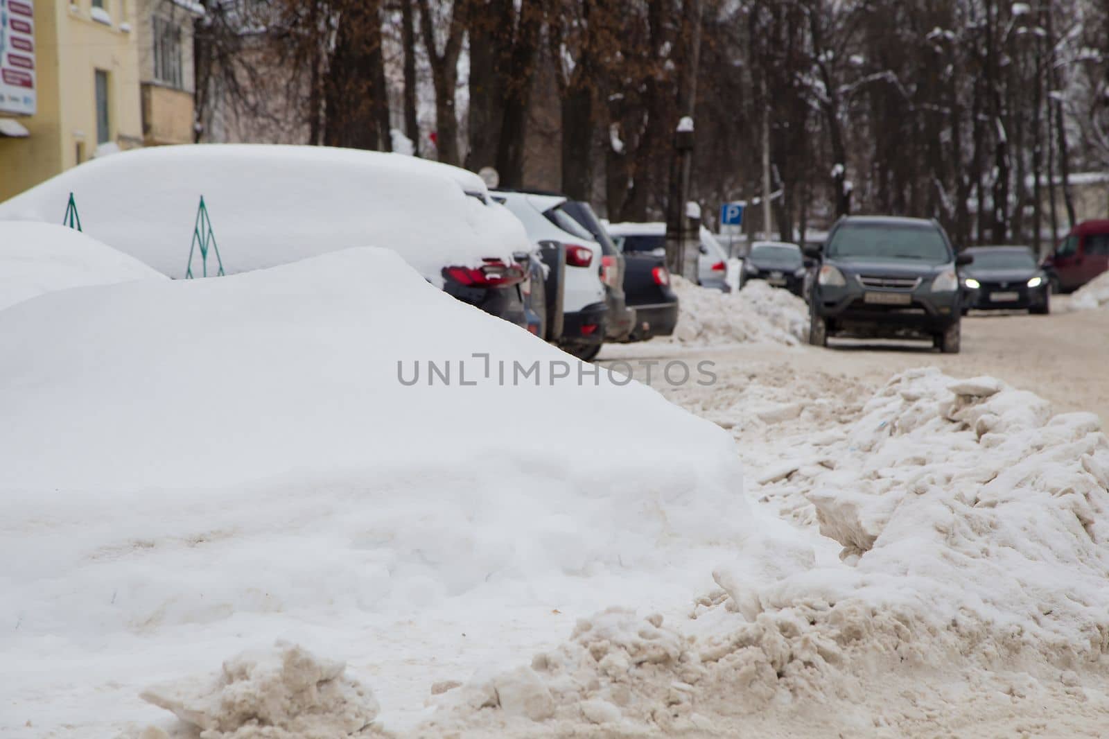 A large snowdrift against the backdrop of a city street with cars. On the road lies white snow in high heaps. Urban winter landscape. Cloudy winter day, soft light.