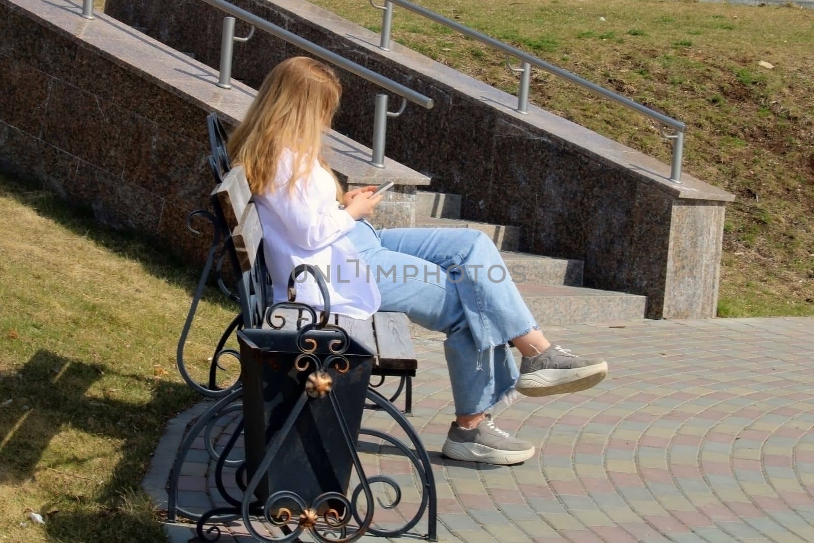 A young blonde with smartphones sits on a bench in the park.