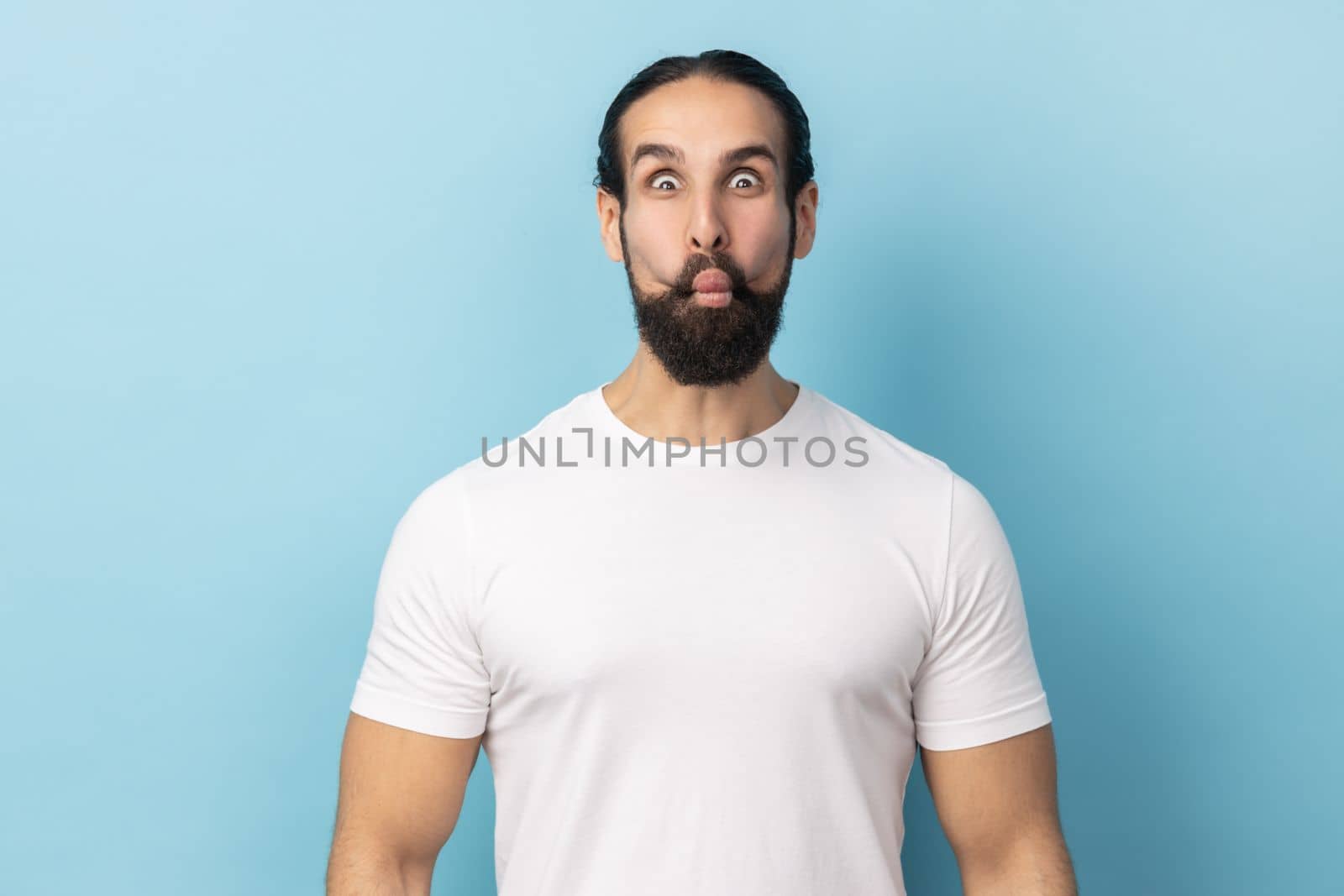 Portrait of childish man with beard wearing white T-shirt making fish face or sending kiss, having fun, kidding, pretending to be stupid. Indoor studio shot isolated on blue background.