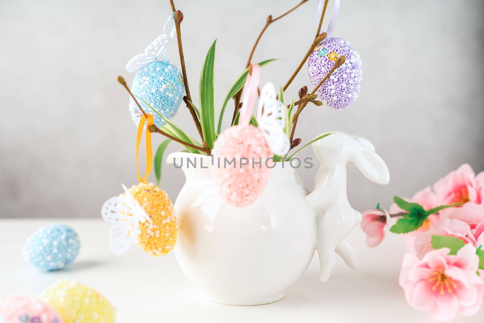 Home interior with easter decor. Vase with willow tree branches with Easter eggs and bunny on white table and background with copy space.