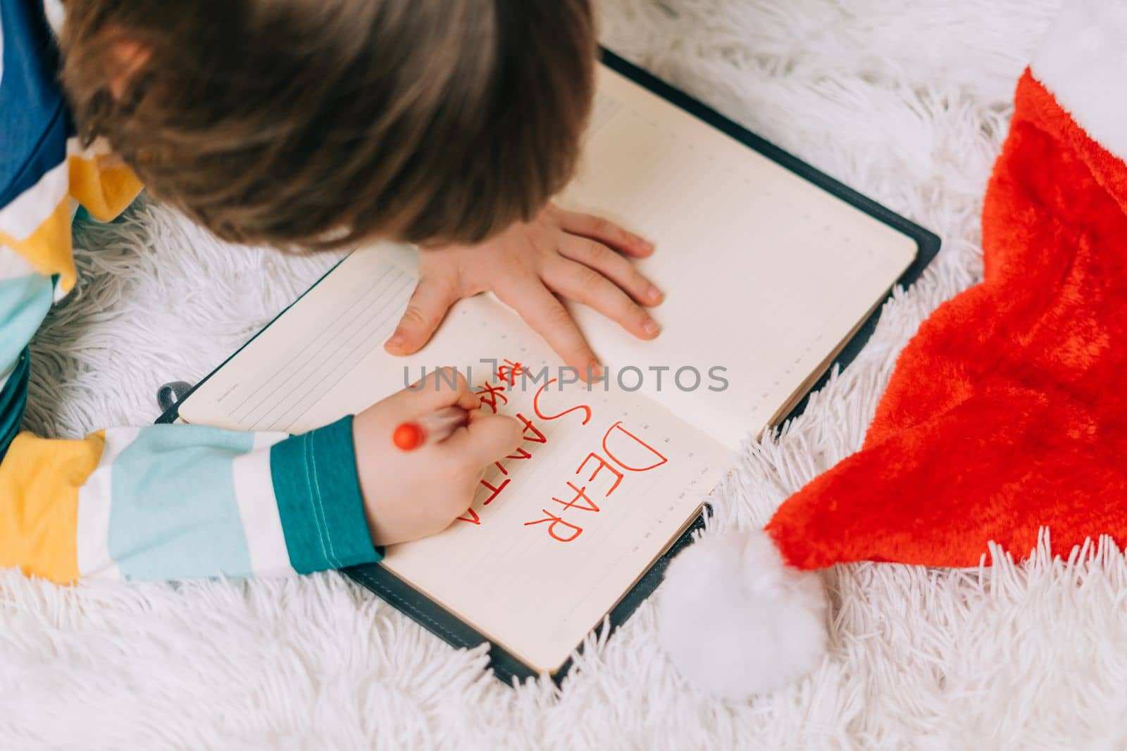 Close up hands of Kid boy, lying on couch and writing letter in note pad to dear Santa at home in Christmas holidays. Child wish list. Dreams of a Christmas gifts. Merry Christmas and Happy new year