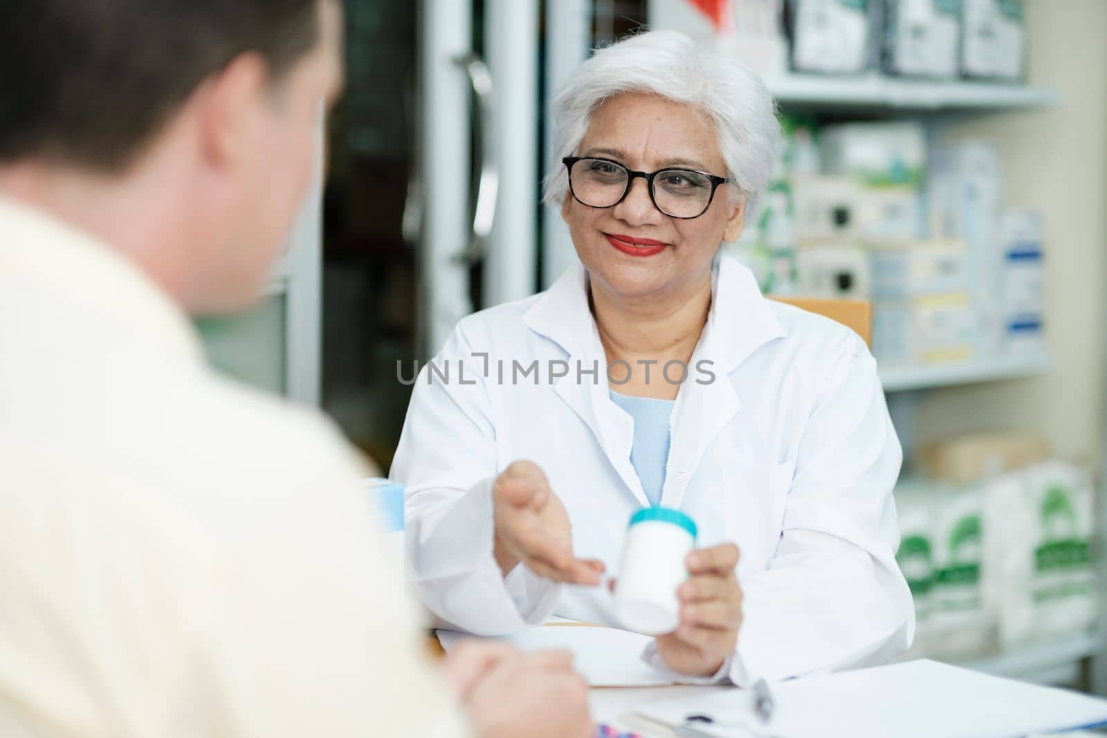 Female senior pharmacist at the drugstore wearing white gown talking, giving advice, explaining, suggesting, and recommending to client or patient about the prescription and medications. Medicine and healthcare concept.