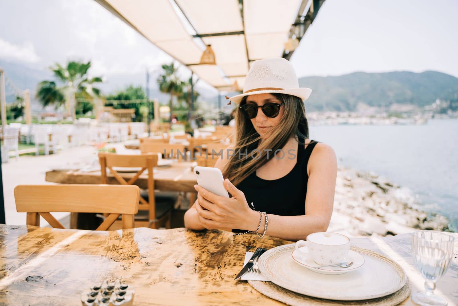 Young girl female lady in sunglasses looking at her mobile smart phone gadget in a street seaside cafe restaurant coffee shop with scenery mountains in the background. Hello summer holiday vacation.