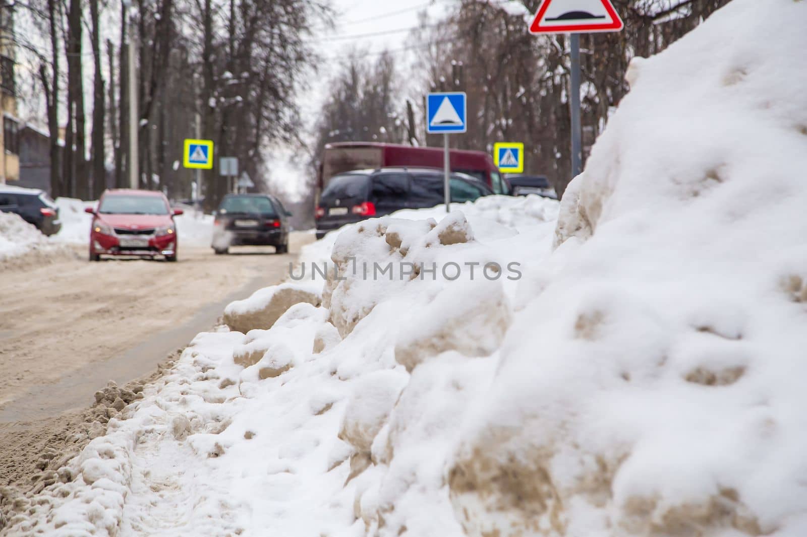 A huge snowdrift against the background of a city street with defocused cars. On the road lies white snow in high heaps. Urban winter landscape. Cloudy winter day, soft light.