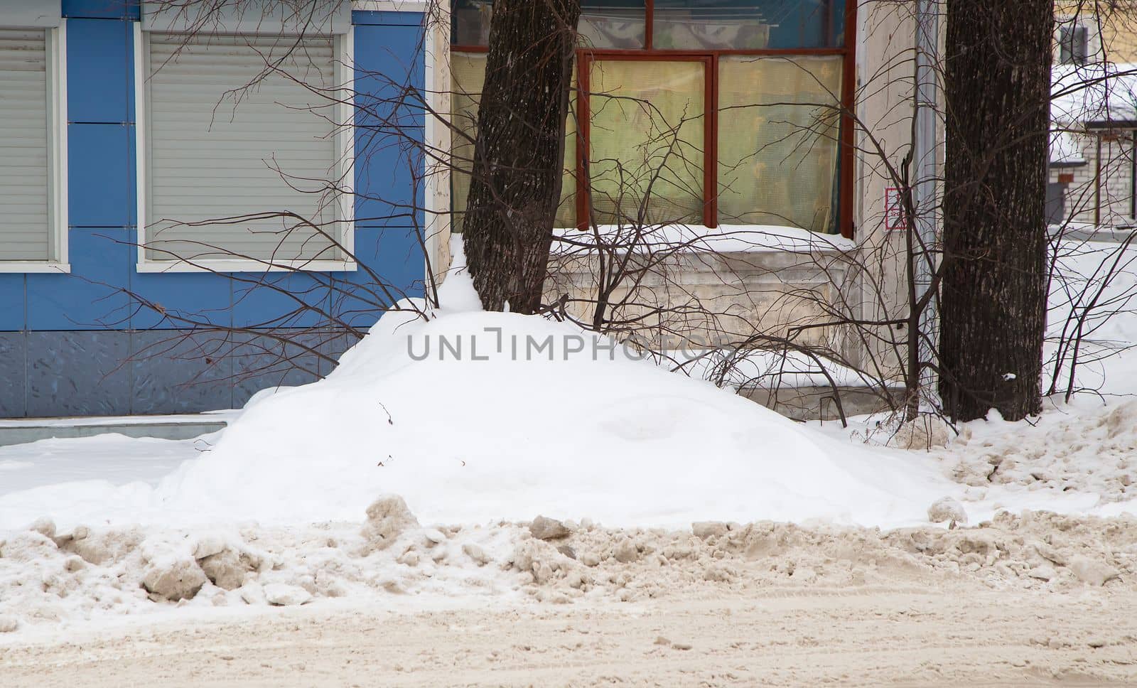 A small snowdrift on the background of the wall of the building. On the road lies white snow in high heaps. Urban winter landscape. Cloudy winter day, soft light.