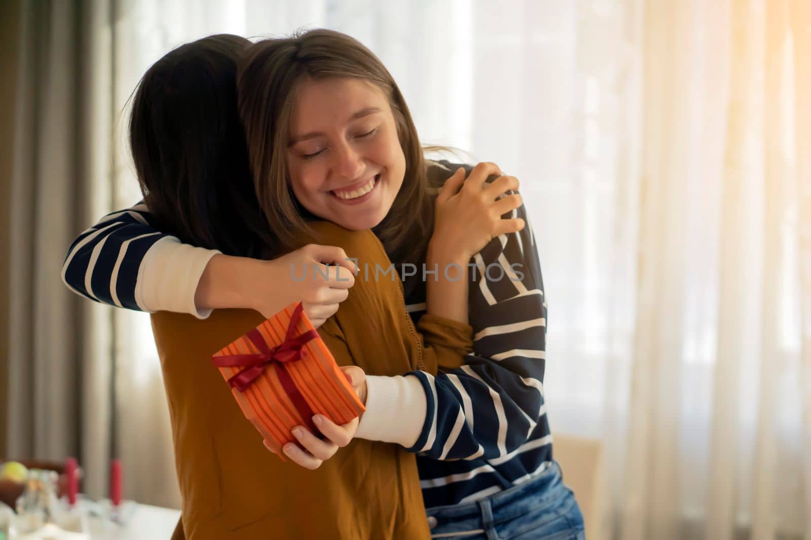 Two happy young girls laugh, friends hug and give each other gifts during the holiday. Women surprise and congratulate on the anniversary of their relationship in their cozy home.