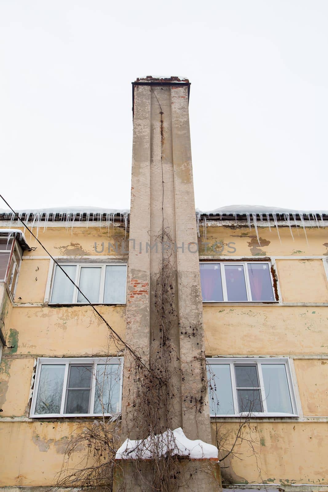 Smoke, stone exhaust pipe along the wall of the old house. There is snow and icicles on the roof, against the background of a gray sky. Cloudy, cold winter day, soft light.