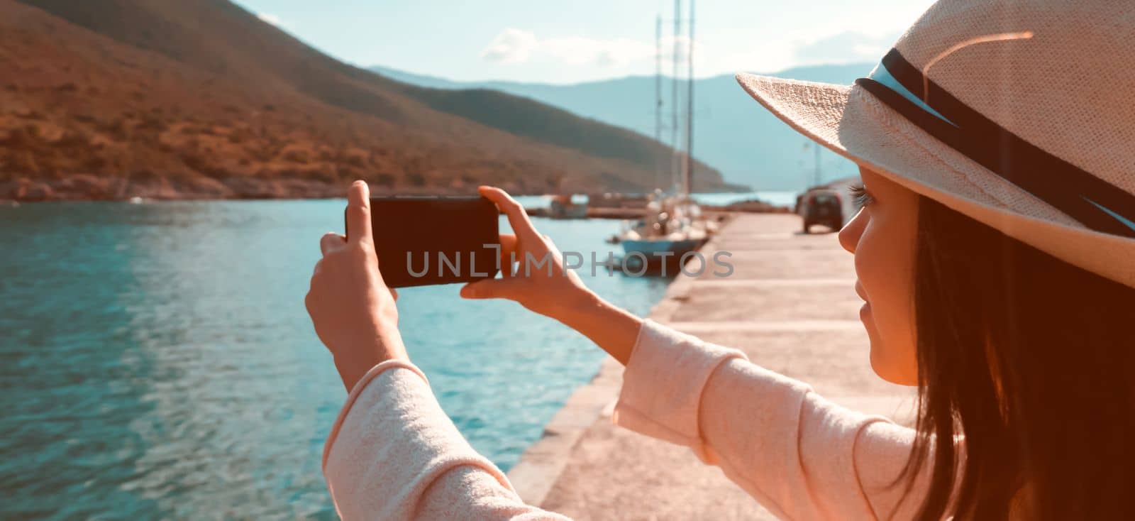 A young girl in a hat and with a backpack makes a photo of a beautiful bay with yachts and boats on the sea while traveling, a tourist walks along the shore next to the ocean and takes pictures.