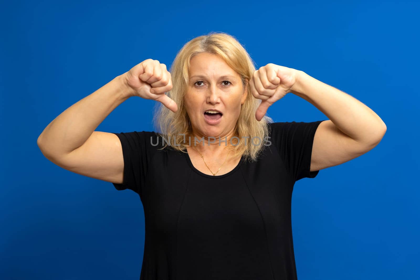 Blonde caucasian woman in her 40s giving thumbs down in disgust isolated on blue studio background