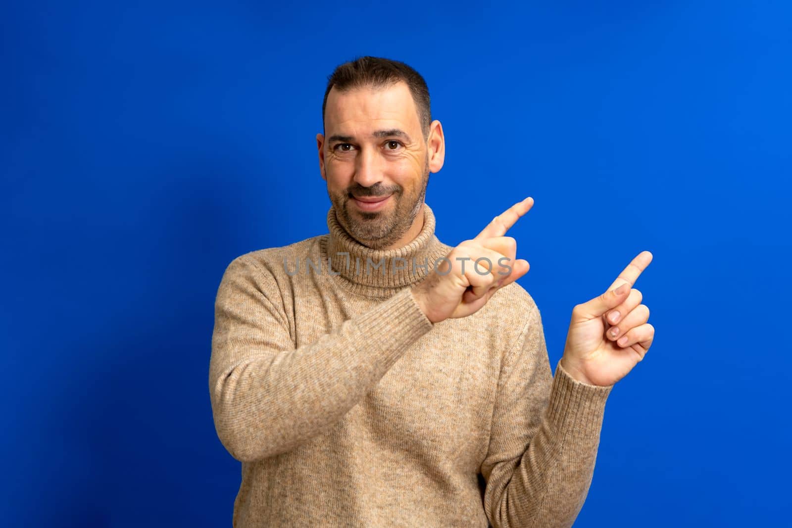 Portrait of a happy latin man in brown turtleneck pointing index fingers at copy space isolated over blue background