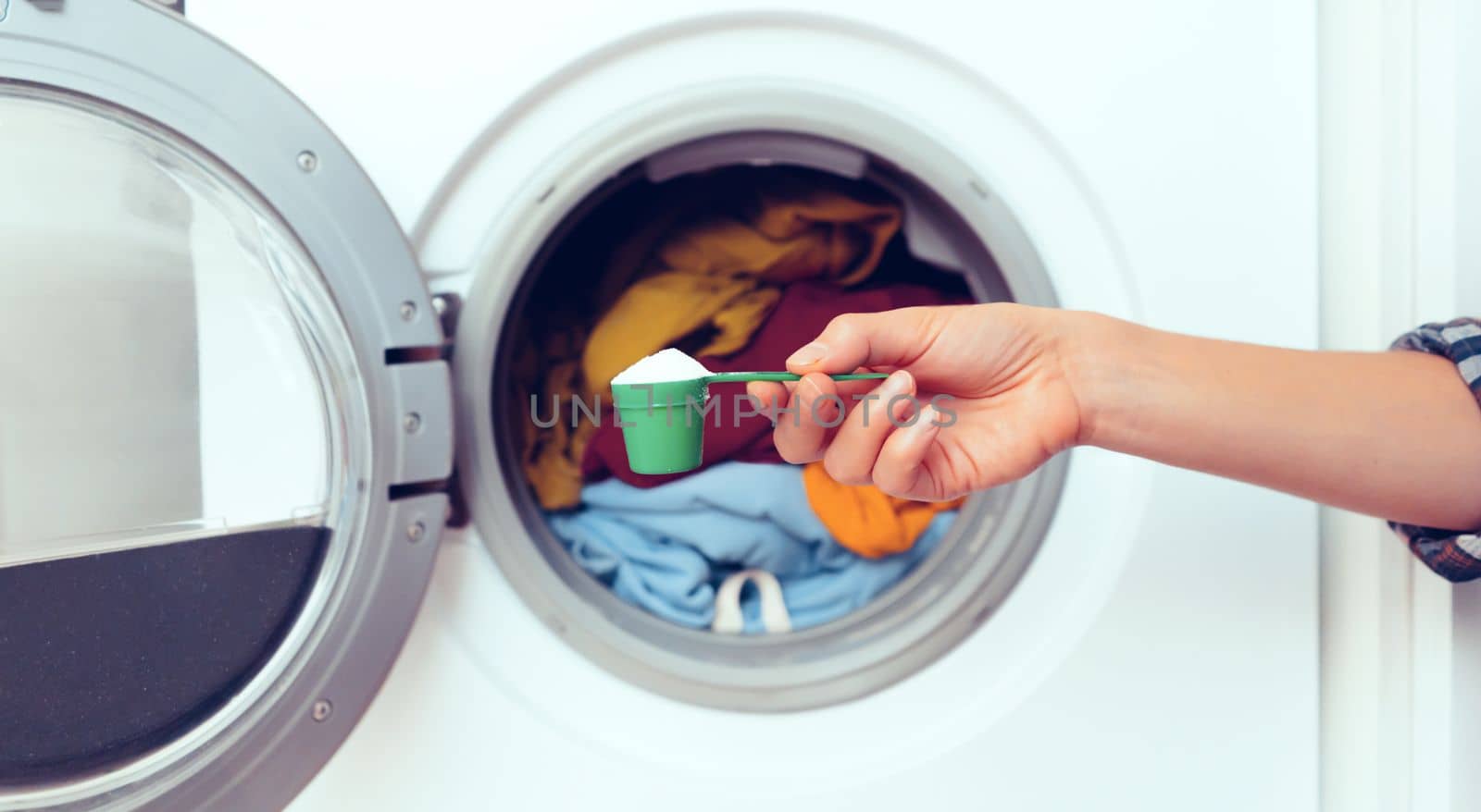 A woman's hand holds a cap with washing powder against the background of a washing machine with bright things put into it. The girl washes and wringes out things in the laundry room at home.