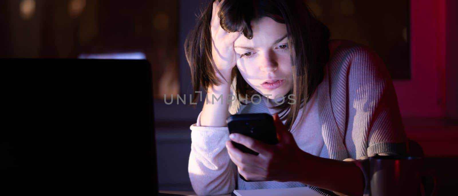 A young girl works at night at home in the dark, uses a laptop and holds her mobile phone in her hand, puzzled and surprised reads a message on her device online, is engaged in a project.