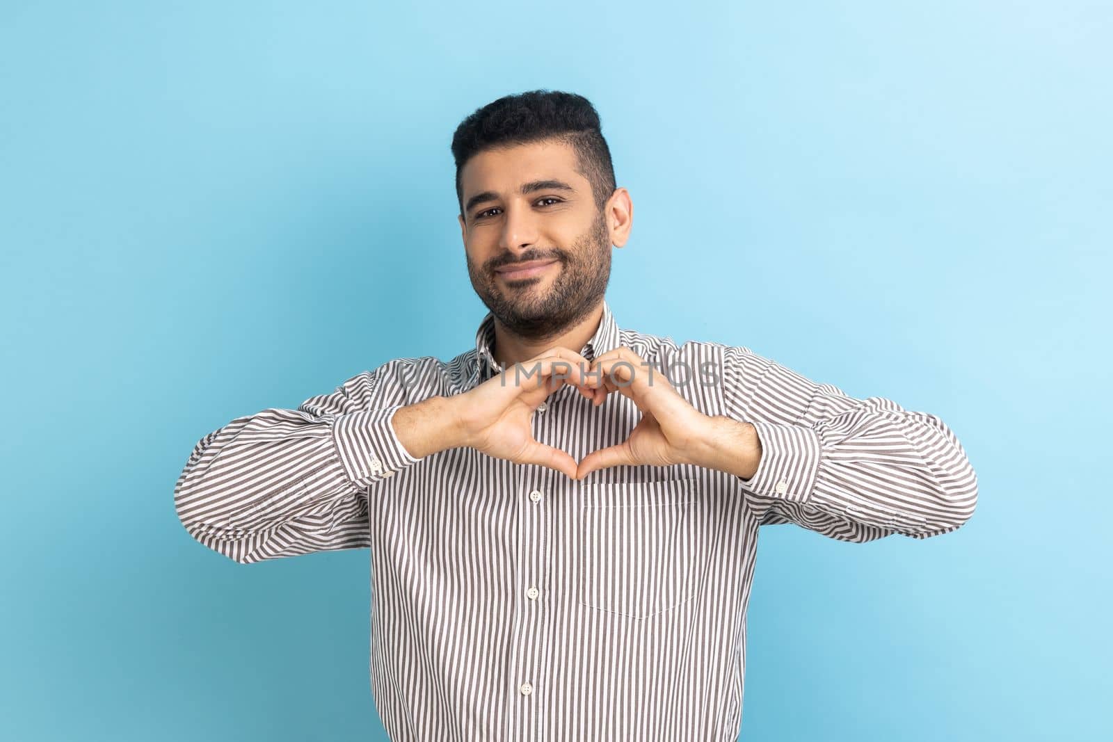 Happy positive handsome businessman with beard standing and showing heart shape making with hands, looking at camera with love, wearing striped shirt. Indoor studio shot isolated on blue background.