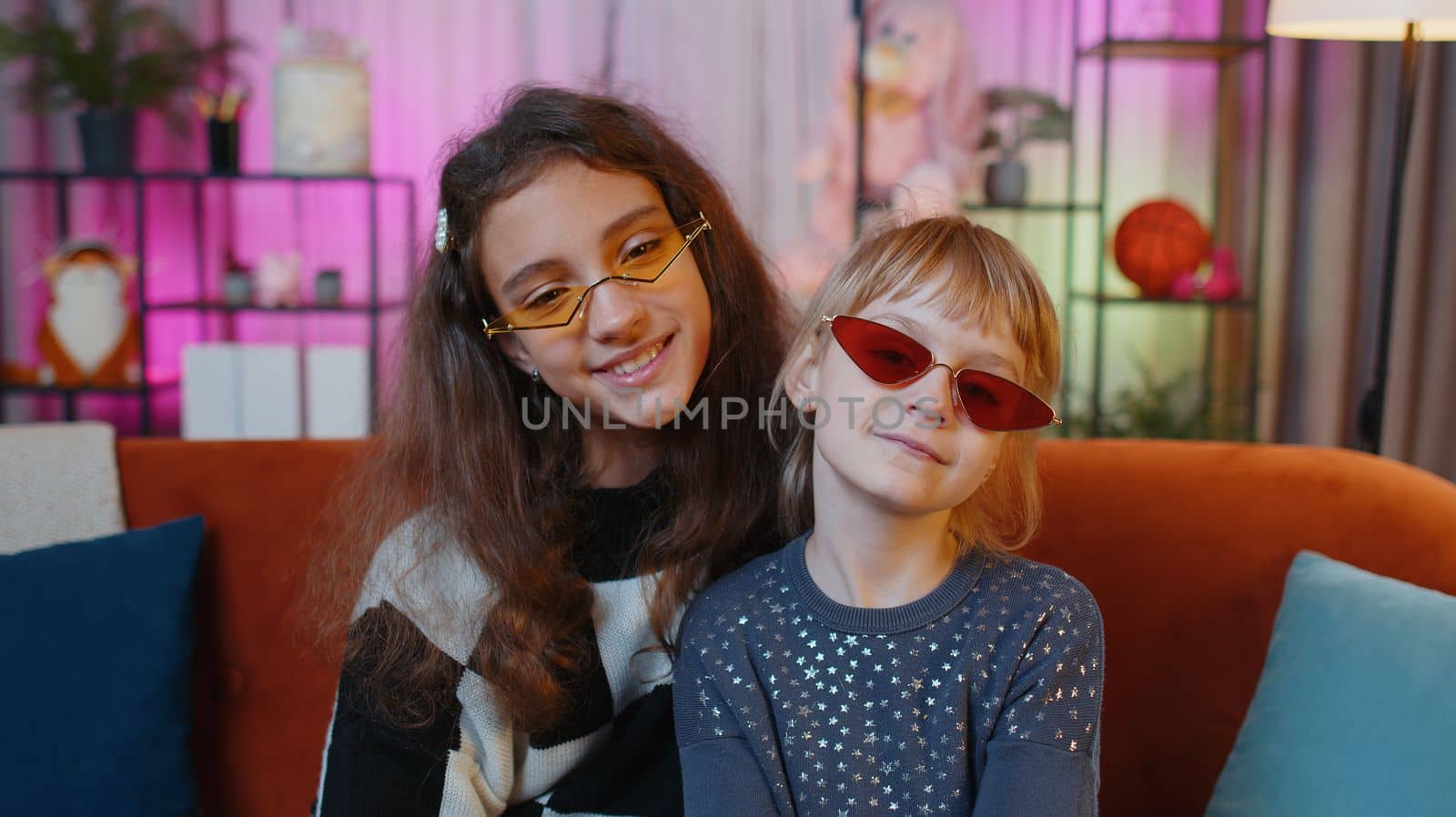 Portrait of happy smiling teenage child and little sister kid looking at camera wearing stylish sunglasses. Female siblings children or best friends sitting on sofa at home playroom. Friendship family