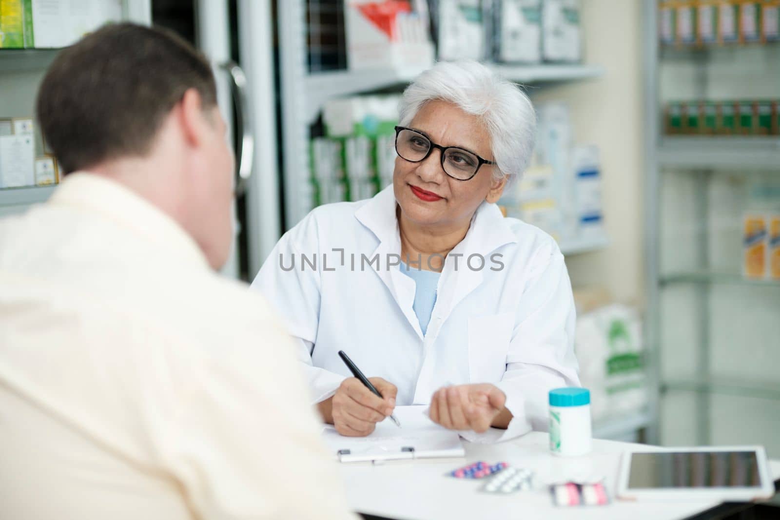 Female senior pharmacist at the drugstore wearing white gown talking, giving advice, explaining, suggesting, and recommending to client or patient about the prescription and medications. Medicine and healthcare concept.