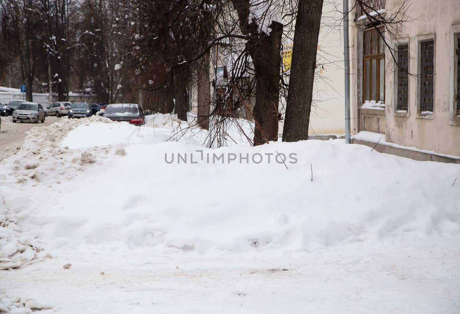 A small snowdrift against the background of a city street with trees. On the road lies white snow in high heaps. Urban winter landscape. Cloudy winter day, soft light.