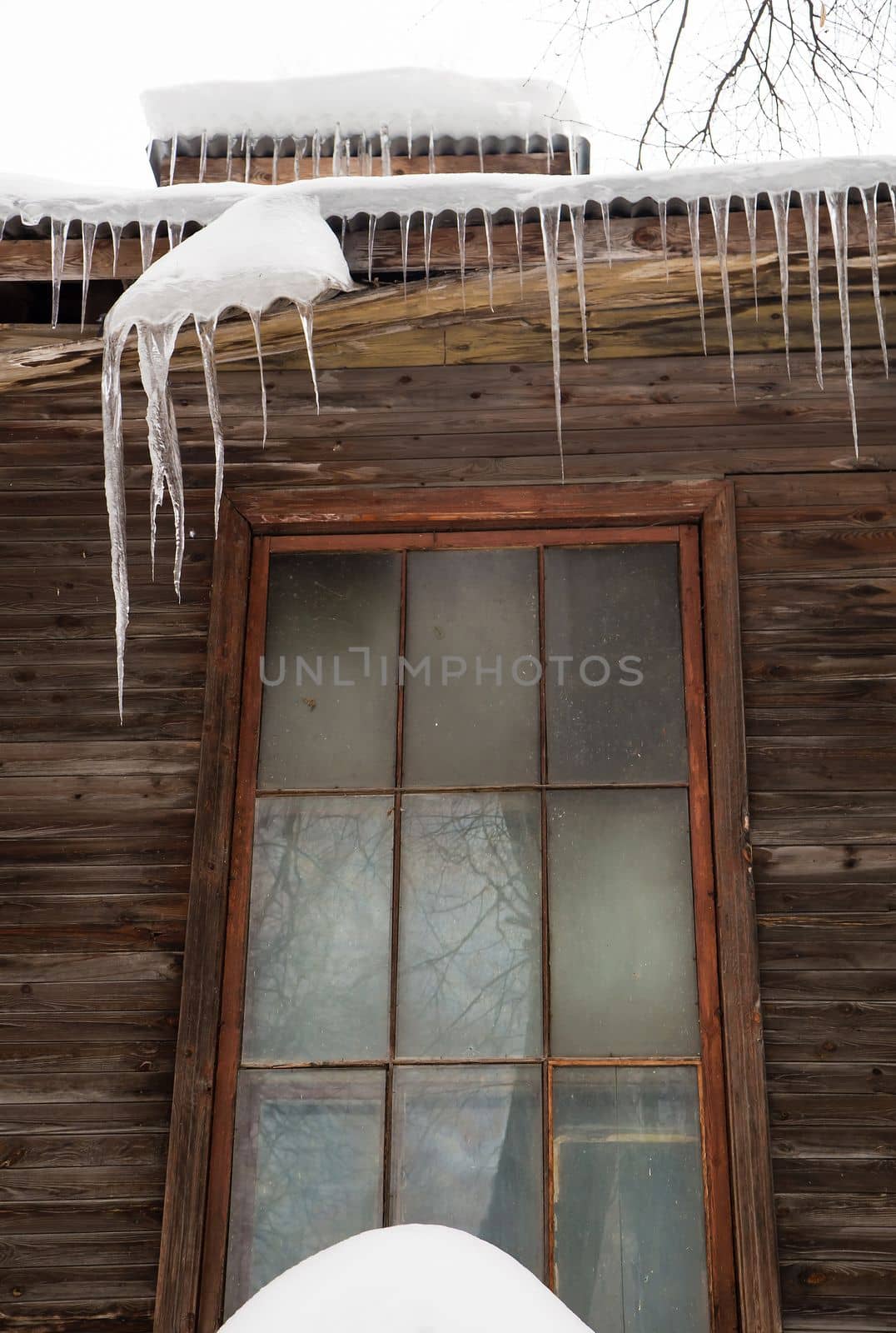 Icy, small icicles hang on the edge of the roof, winter or spring. Plank wall of an old wooden house with windows. Large cascades of icicles in smooth, beautiful rows. Cloudy winter day, soft light.