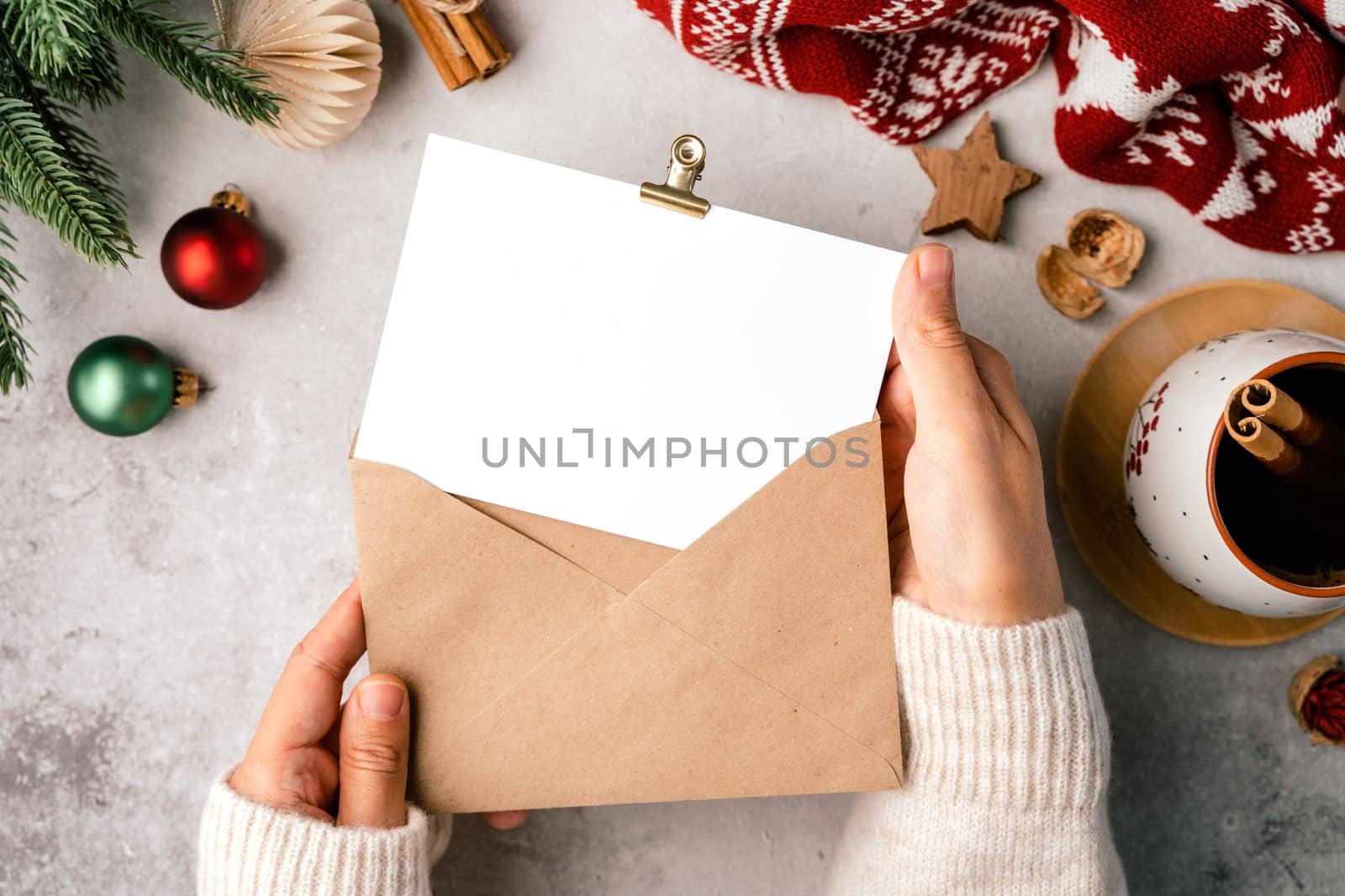 Woman hands in sweater holding blank paper and envelope . Flat lay of gray background with cup of coffee and Christmas decoration. Top view mock up and copy space for text.