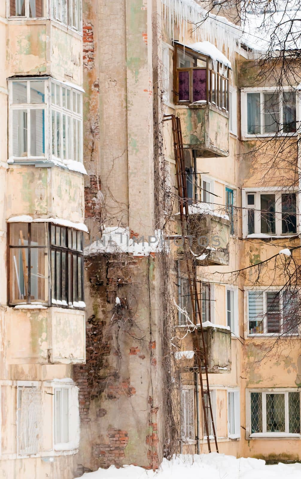 An old four-story house with glazed balconies, a stone chimney on the wall. Plastic windows and an iron ladder on the wall of a shabby house. Cloudy winter day, soft light.