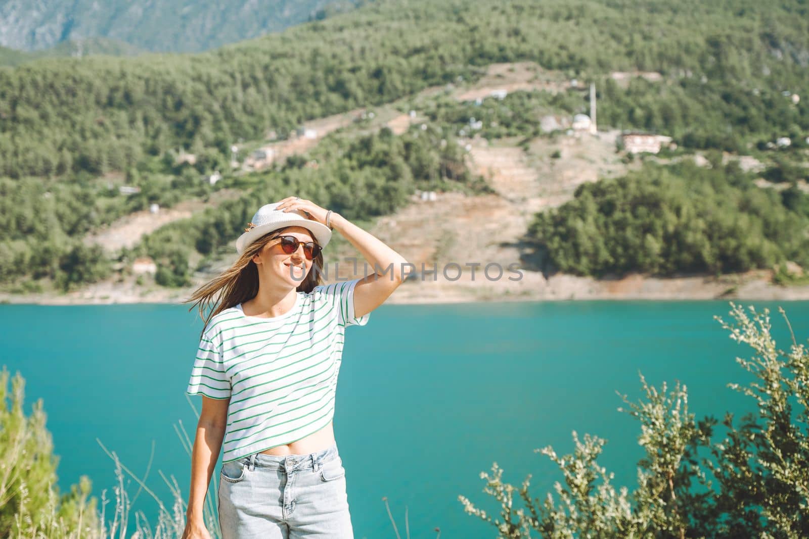 Smiling woman in hat and sunglasses with wild hair standing near mountains lake on background. Positive young woman traveling on blue lake outdoors travel adventure vacation.