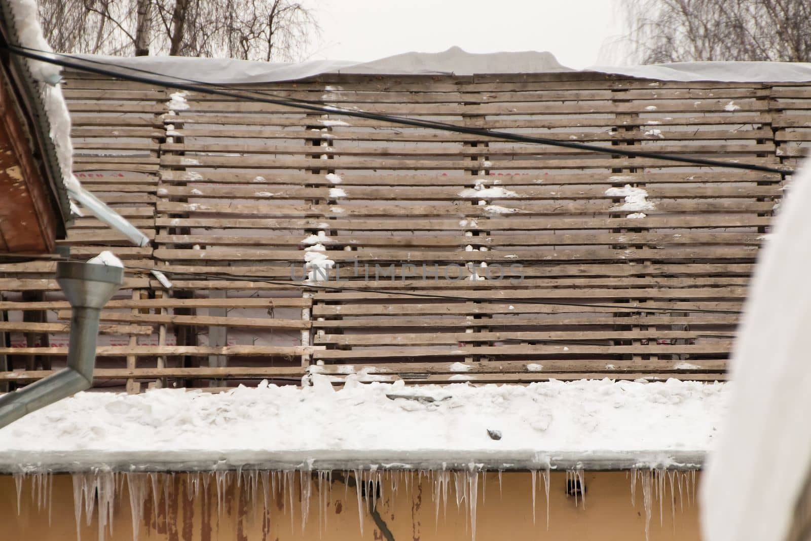 A dismantled roof with a wooden crate and hanging icicles on the edge. An old house against the backdrop of a gray sky. Large cascades of icicles hang in a row. Cloudy winter day, soft light.