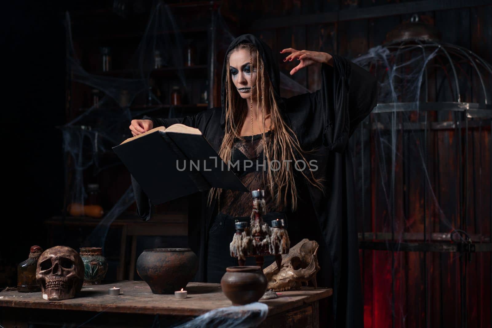Witch dressed dark dungeon room use magic book for conjuring magic spell. Female necromancer wizard gothic interior with skull, cage, spider web