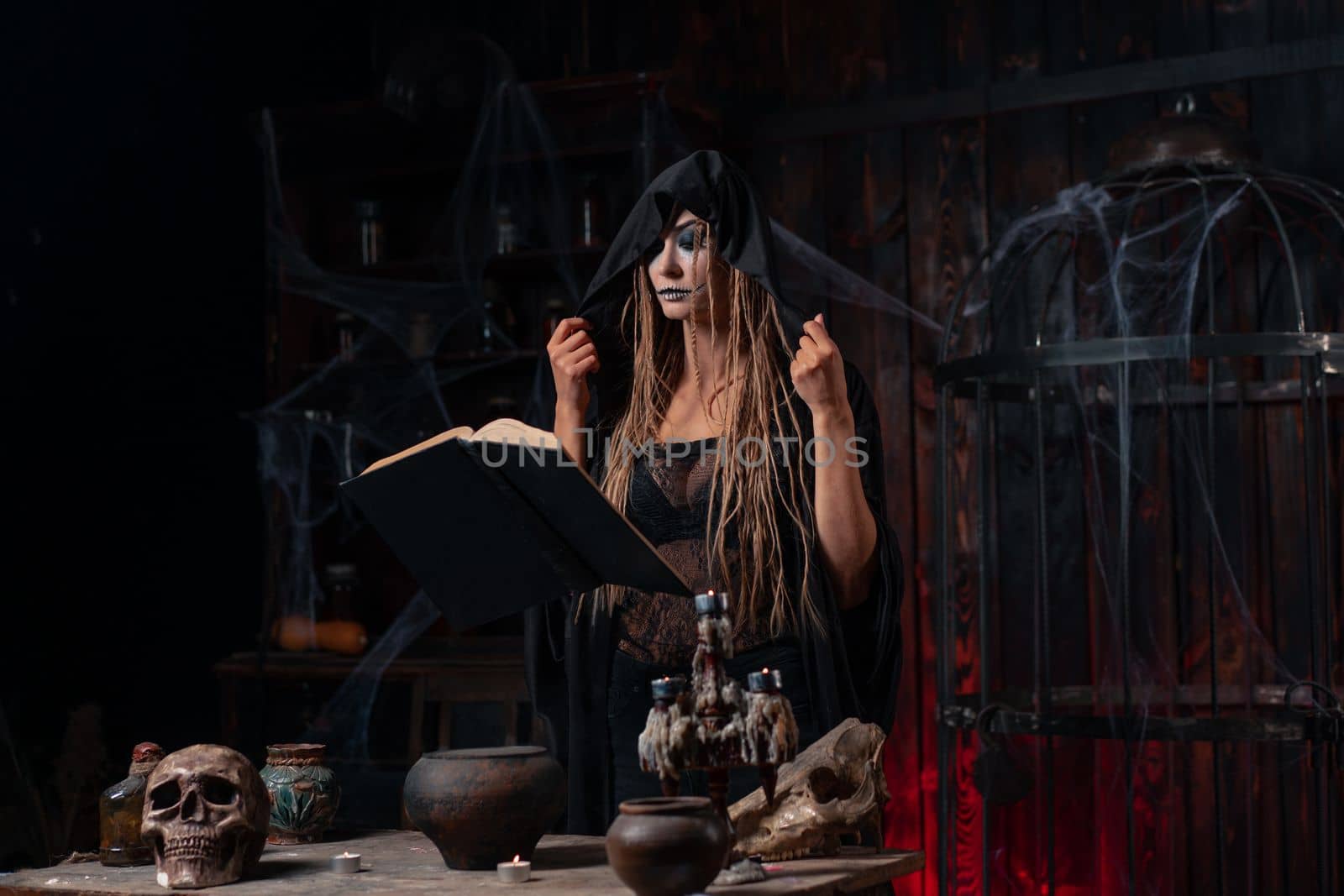 Witch dressed black hood with dreadlocks standing dark dungeon room use magic book for conjuring magic spell. Female necromancer occultist medium shoot