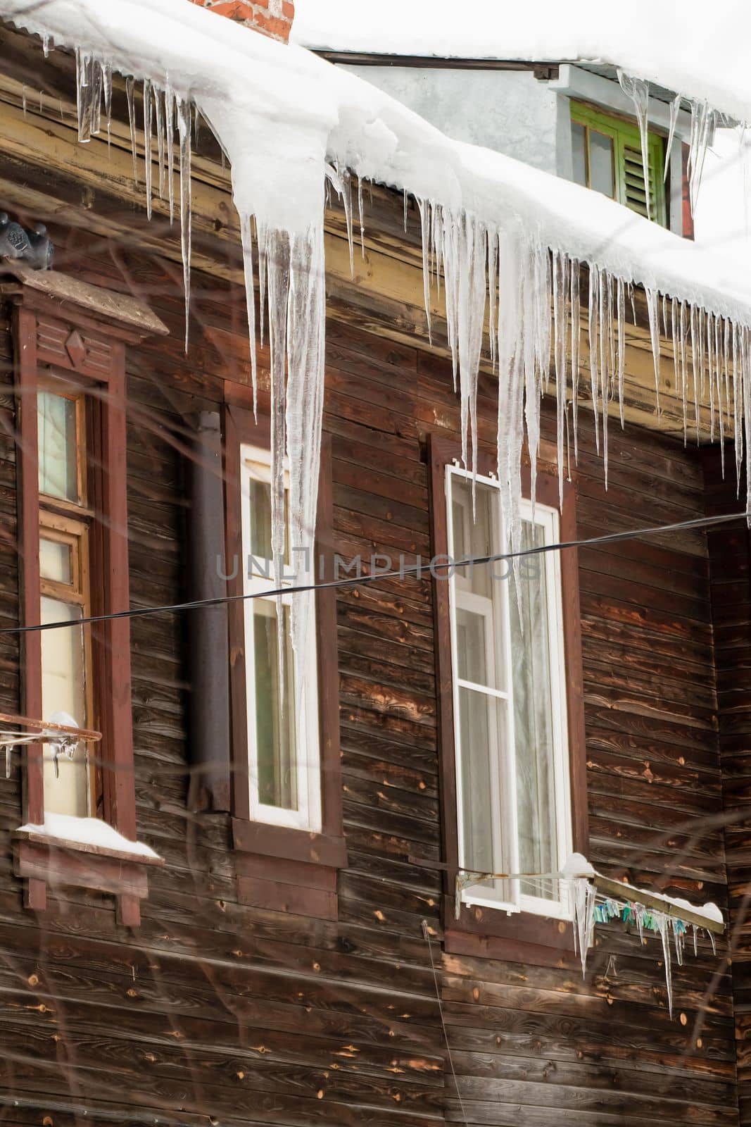 Multiple icicles hang on the edge of the roof, winter or spring. Log wall of an old wooden house with windows. Large cascades of icicles in smooth, beautiful rows. Cloudy winter day, soft light.